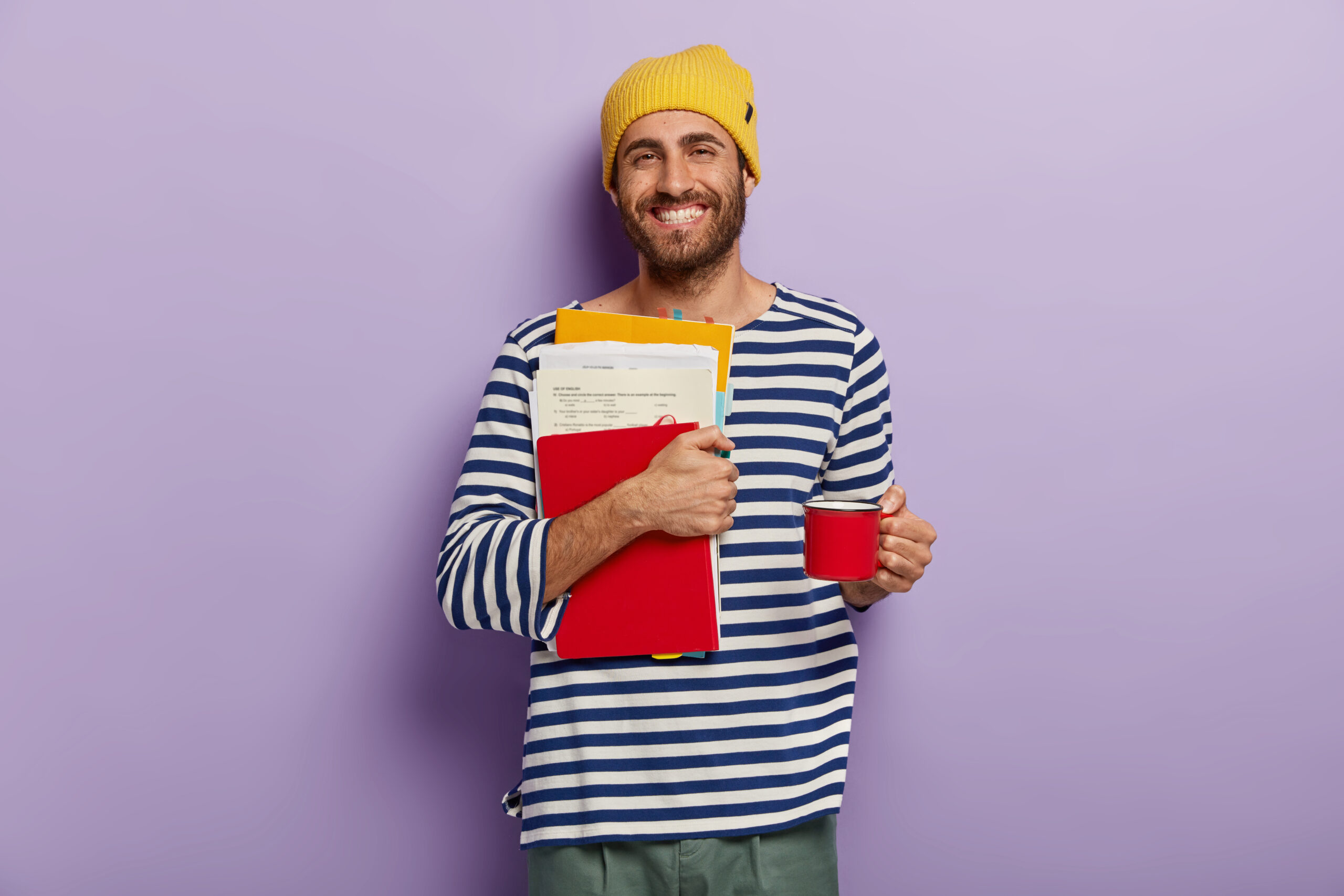 happy college student carries papers and notepad, prepares for exams, drinks hot beverage from cup, wears striped clothing, isolated over purple wall, studies at high school. education concept