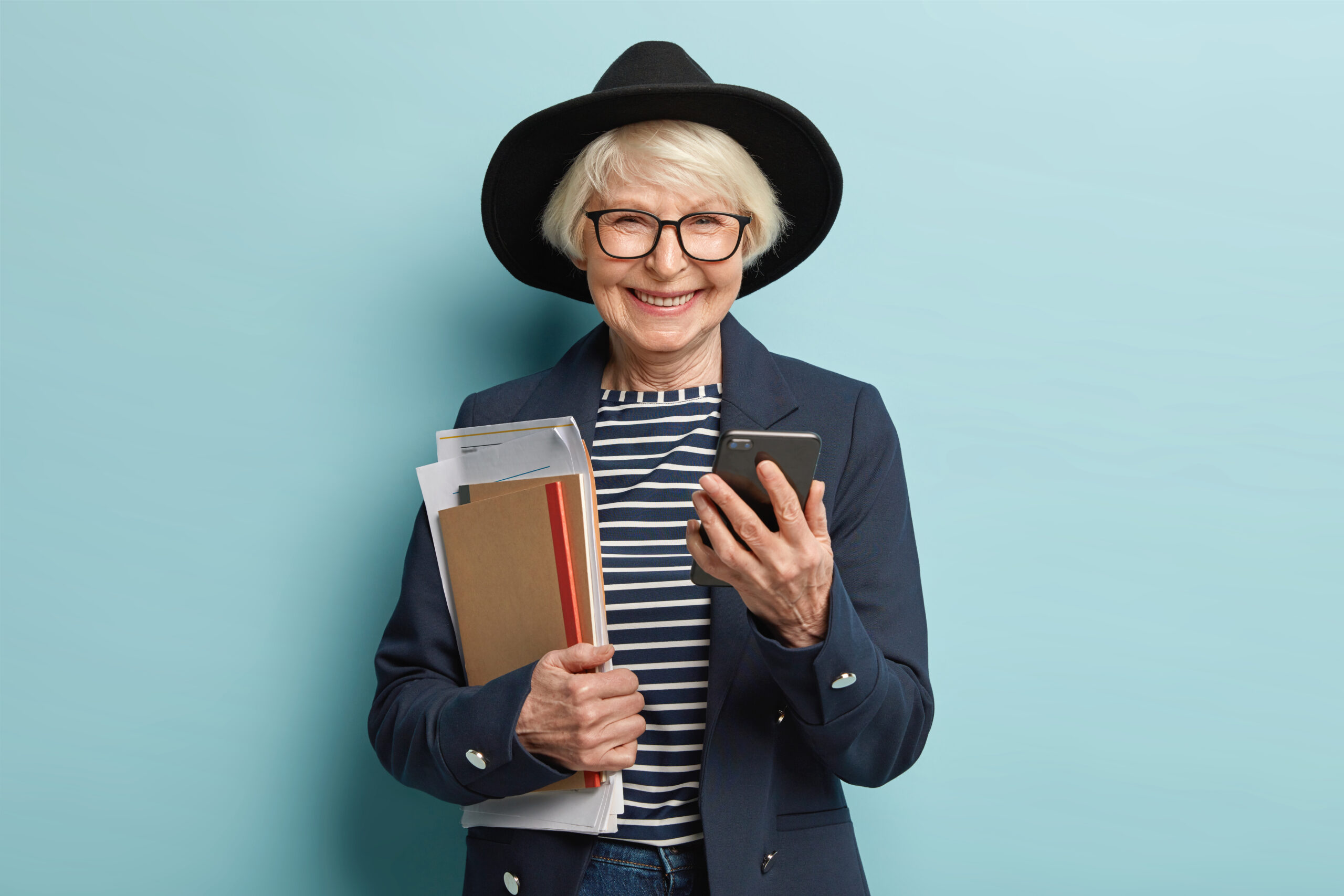 successful elderly smiling businesswoman checks data information on mobile phone, holds notepad with documents, returns from important conference, dressed in stylish clothes, makes payment online